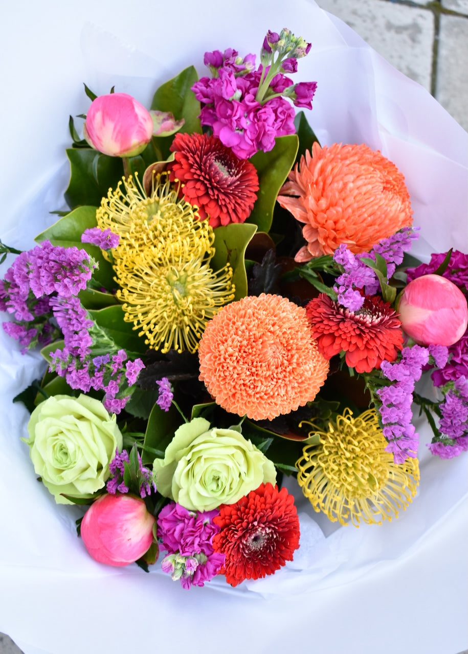 Bright & colourful fresh flower bouquet. Created in colours of pink, bright orange, red & lime green. Crafted by a florist & presented in a white paper wrap.
