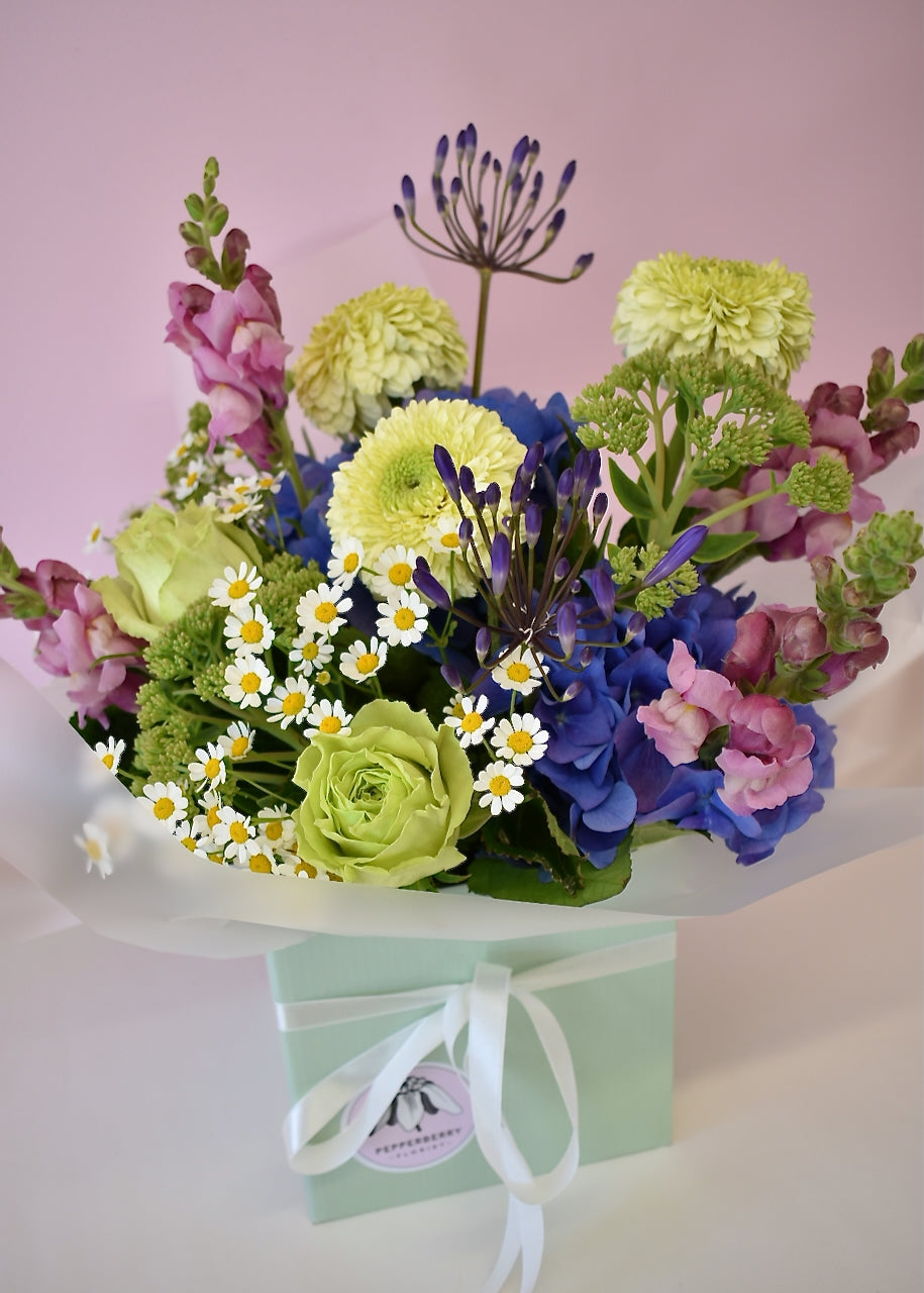 A colorful floral arrangement featuring various flowers including daisies and roses, in a cardboard box with a ribbon.