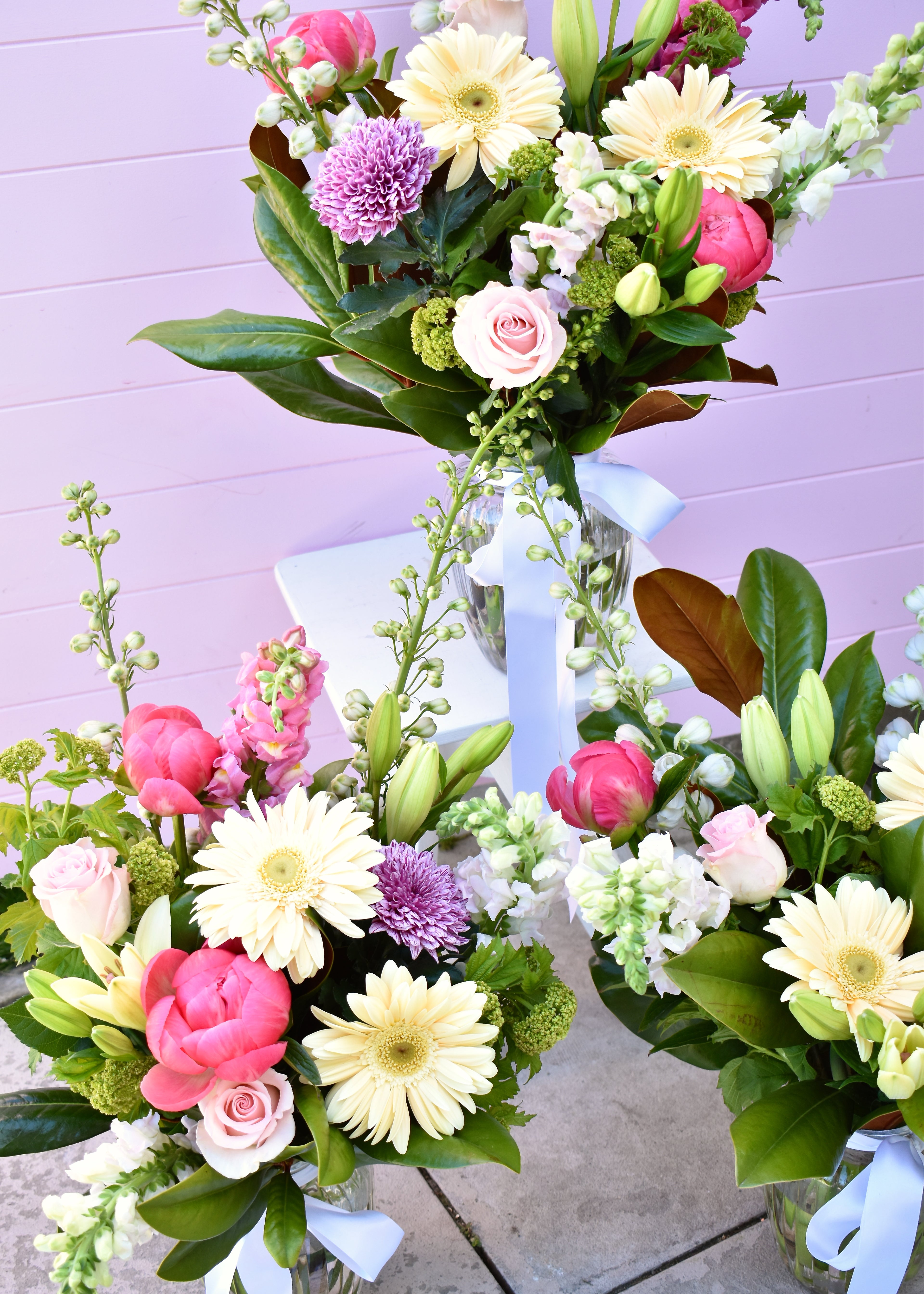 A bouquet of pastel-colored flowers including pink roses, gerbera daisies, and various greenery, arranged and presented in a glass vase, set against a pink background.