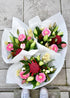 Three fresh flower bouquets wrapped in white paper. Created in pink and white tones. Flowers include gerberas, tulips, freesias and lilies with foliage.