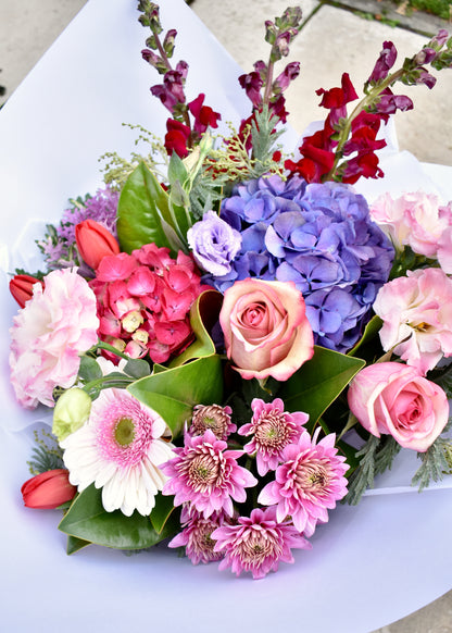 Berry coloured fresh flower bouquet. Red, pink and white. Blue hydrangea, pink roses and mauve chrysanthemums. In white paper wrap.