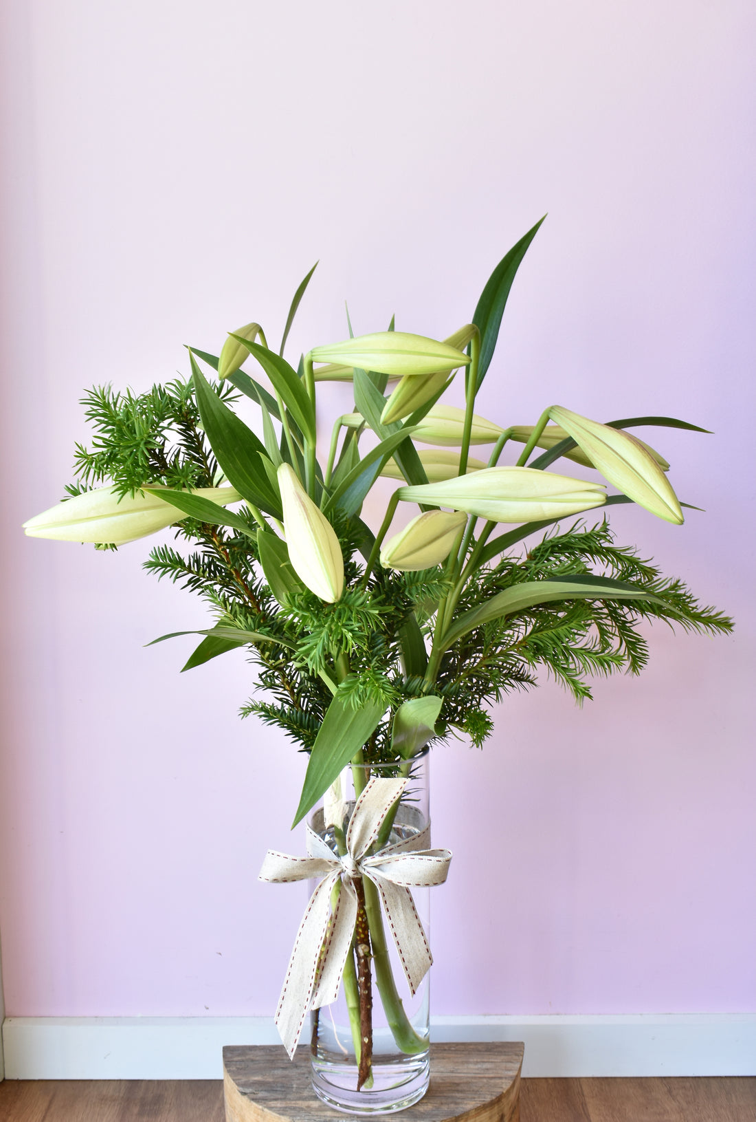 Bouquet of white Christmas lilies with festive greenery in a glass cylinder vase.