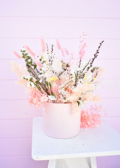 Dried flower arrangement sitting on a white stool. A blush pink pot containing a flower arrangement in pink, blush, peach and white