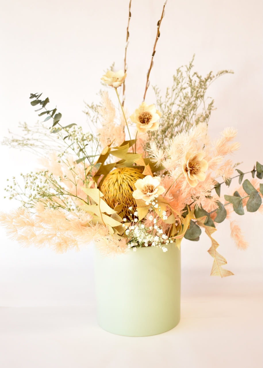 A soft pastel green ceramic pot containing a wild flower dried flower arrangement. Created in colours of mustard, cream, peach and white and made with fern, banksia, gypsophlia, statice, eucyalyptus and hand made wooden roses.