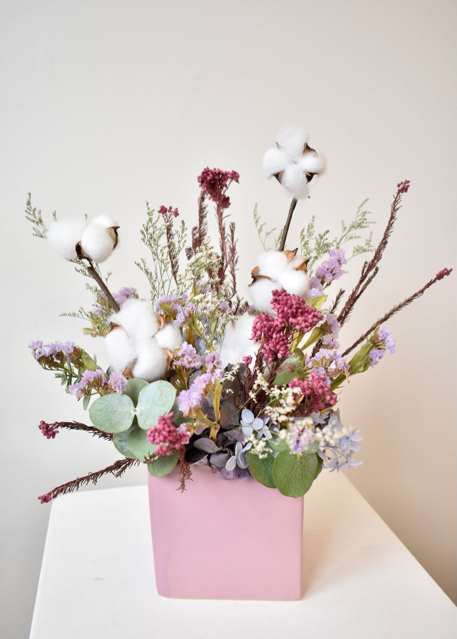 Ceramic mauve coloured square vase filled with a dried flower arrangement. Cotton flower, hydrangea, rice flower and eucalyptus. Colours of white, pale blue, mauve and dusty deep pink.