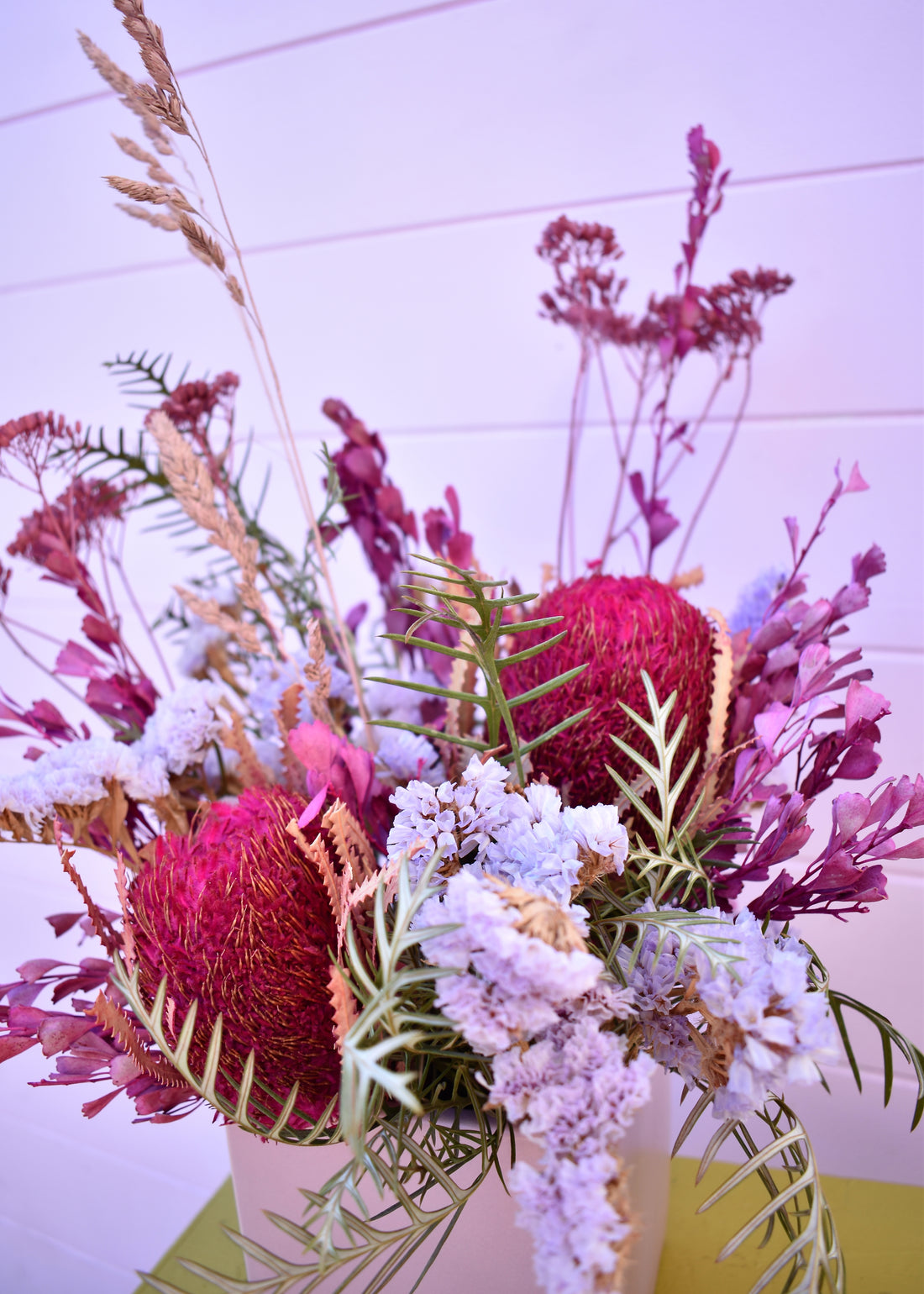 Colorful dried floral arrangement with pink, purple, and green flowers in a purple ceramic vase on a light purple background.