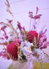 Colorful dried floral arrangement with pink, purple, and green flowers in a purple ceramic vase on a light purple background.