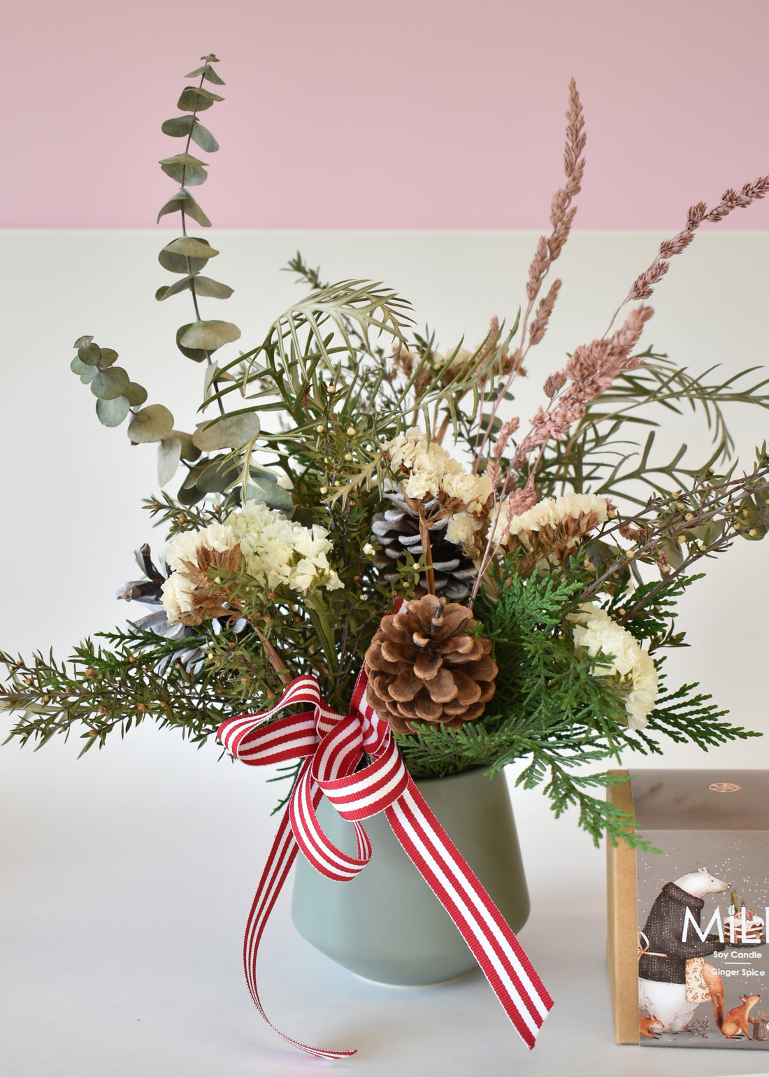 Decorative flower arrangement with pine cones and a striped ribbon in a green vase on a light pink background.