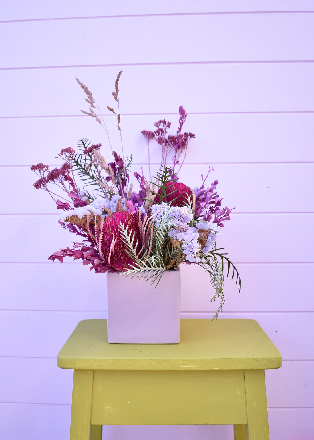 Colourful dried floral arrangement in a pink vase on a yellow stool against a light purple background