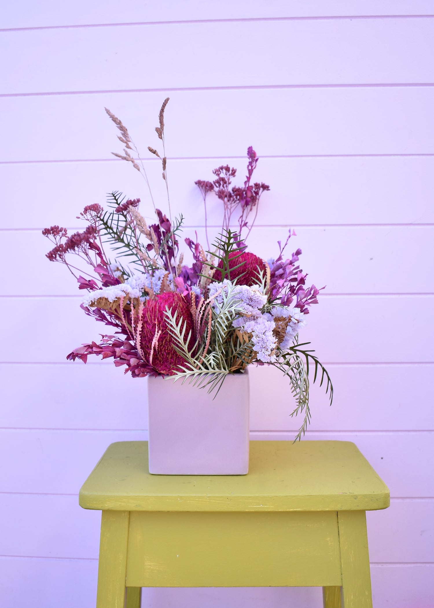Colourful dried floral arrangement in a pink vase on a yellow stool against a light purple background