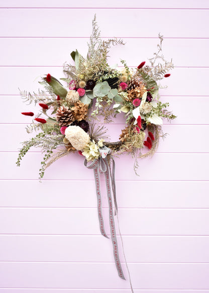 A Christmas wreath made with dried and everlasting flowers. Banksia, eucalyptus, strawflowers, fern and Christmas ribbon. Made by a Florist and on a pink background.