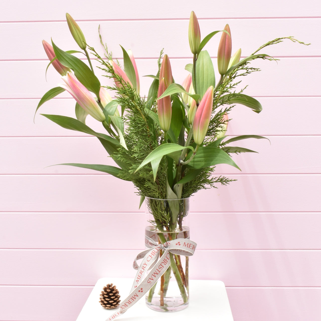 A glass vase of pink oriental lilies with winter Christmas foliage &amp; Christmas with a Merry Christmas fabric ribbon