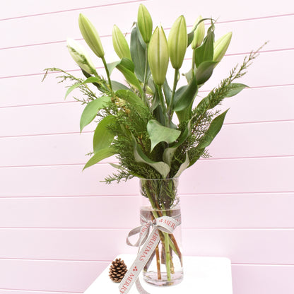 White Oriental Christmas lilies in a glass vase with Christmas foliage and Christmas ribbon