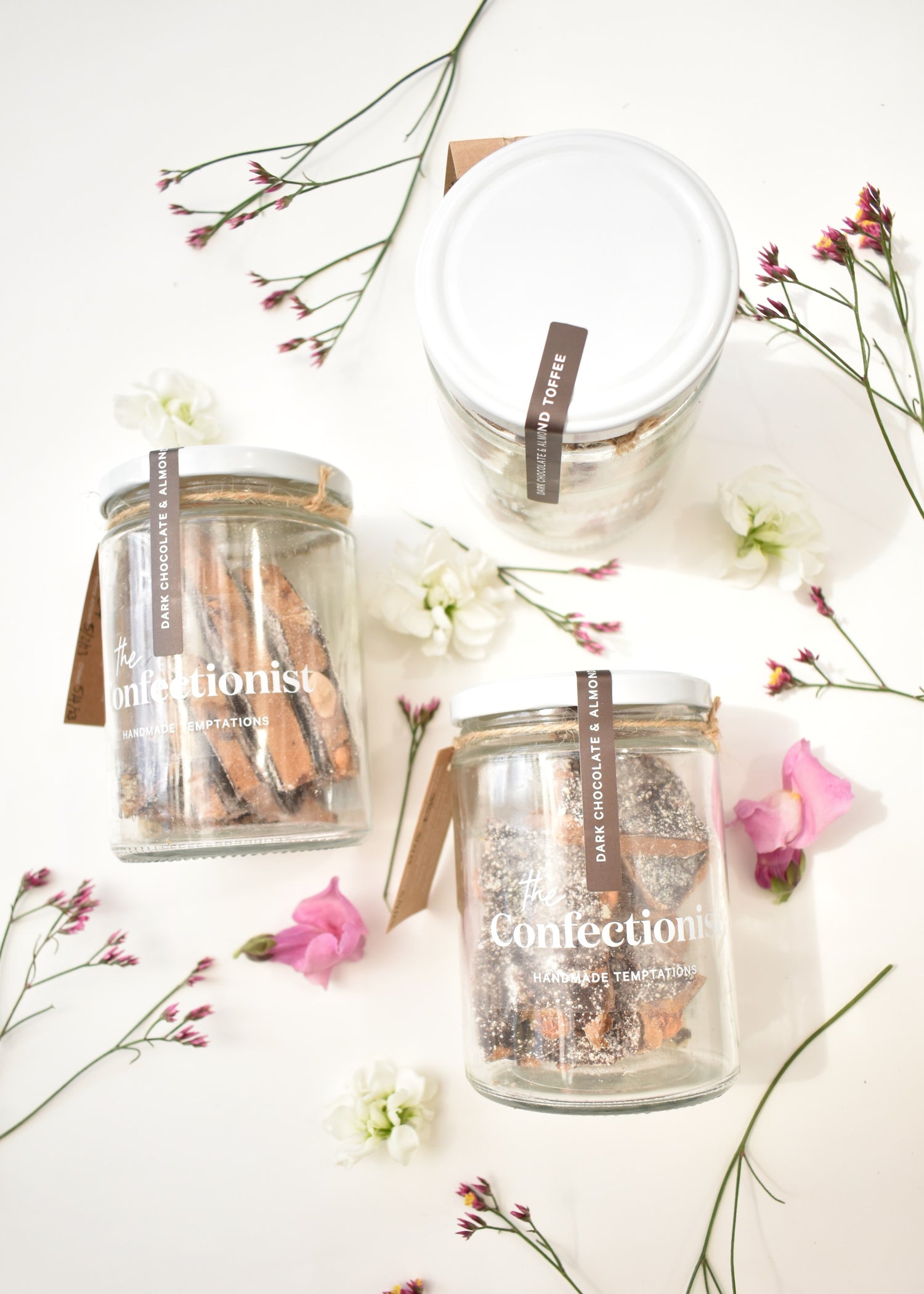 Flat lay image of jars of dark chocolate and almond toffee. White background with pink and white flowers