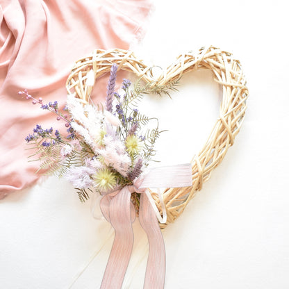A cream wicker heart wreath on a pink backdrop, with a posy of dried flowers in white, cream, pink and purple with pale pink ribbon