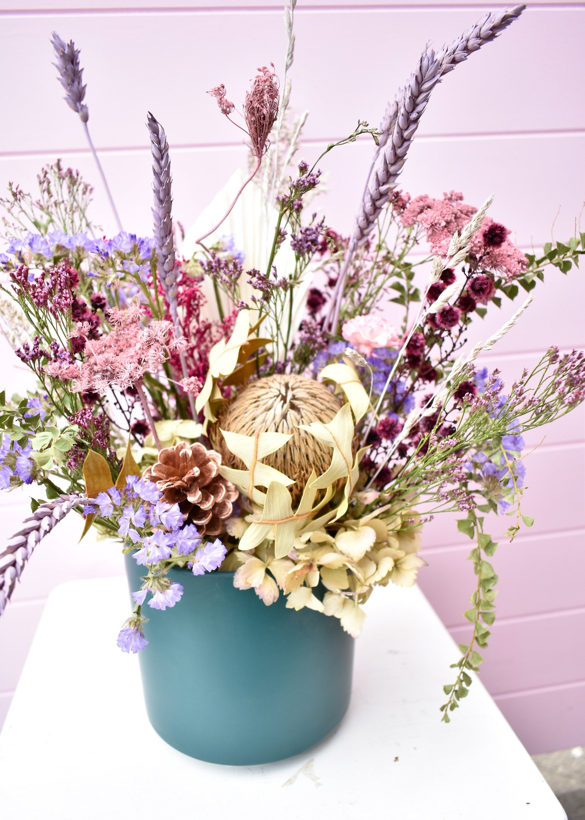 Pink background with a dried flower arrangement sitting on a white stool. A teal green ceramic pot with sage green, lilac, burgundy and lavender everlasting and dried flowers