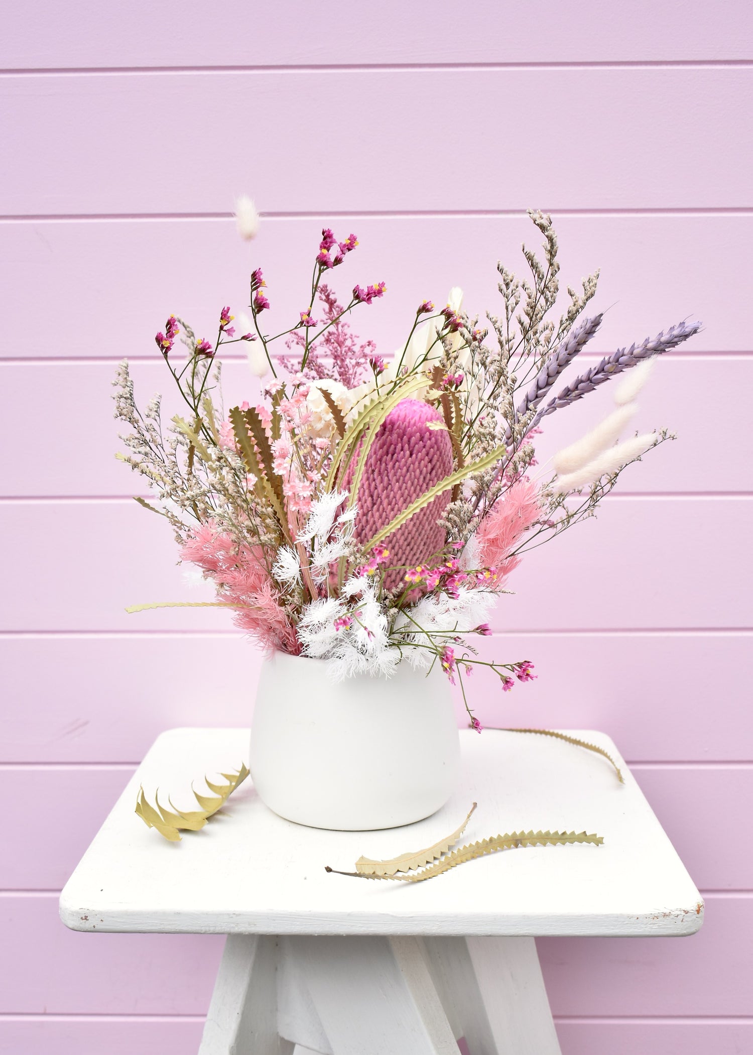 Dried flower arrangement in a white ceramic pot. Pink and white everlasting flowers, banksia, wheat, misty, ming fern, bunny tails and more. Sitting on a white stool in front of a pale pink backdrop.