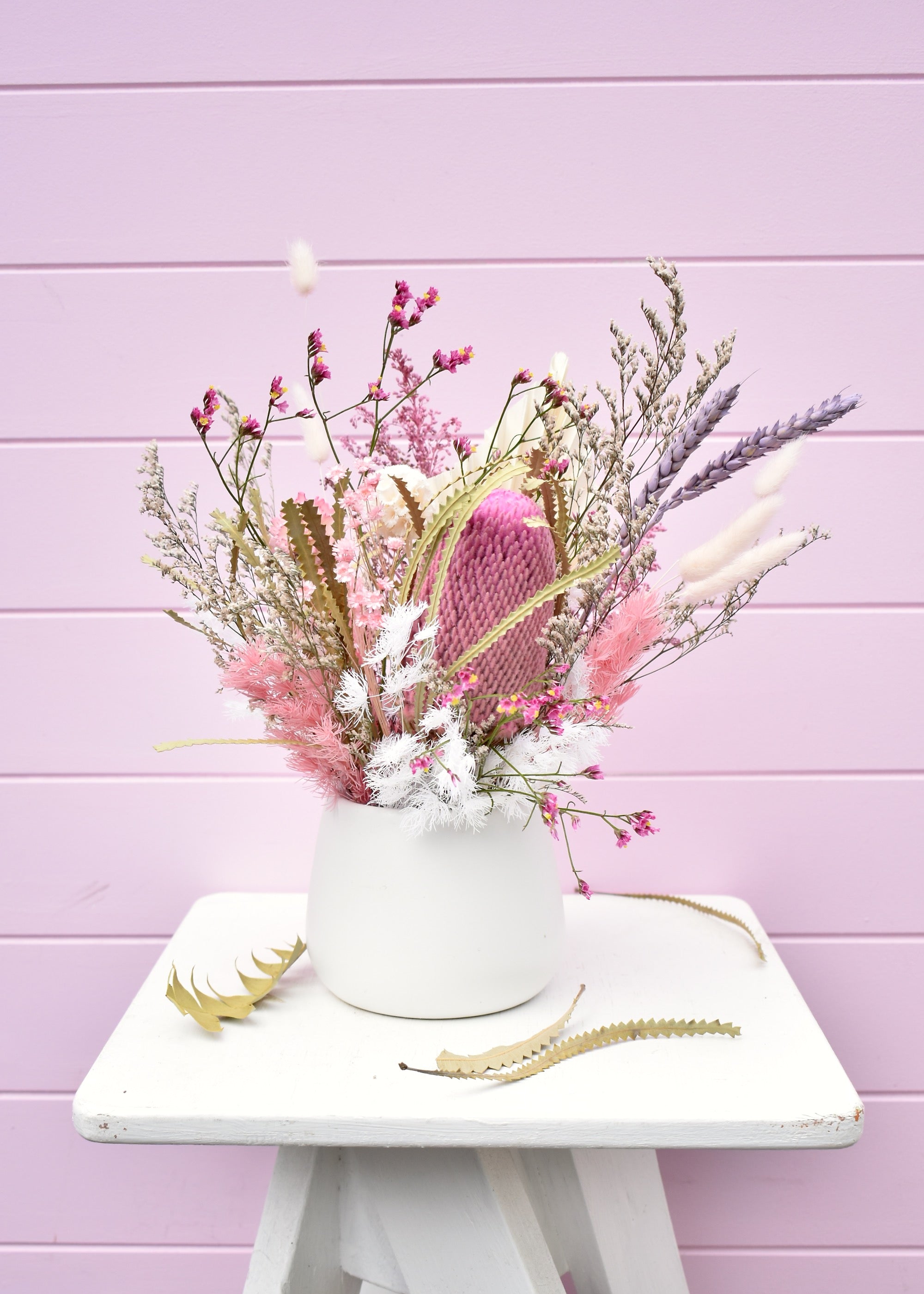 Dried flower arrangement in a white ceramic pot. Pink and white everlasting flowers, banksia, wheat, misty, ming fern, bunny tails and more. Sitting on a white stool in front of a pale pink backdrop.