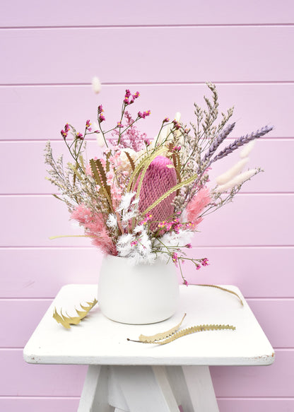 Dried flower arrangement in a white ceramic pot. Pink and white everlasting flowers, banksia, wheat, misty, ming fern, bunny tails and more. Sitting on a white stool in front of a pale pink backdrop.