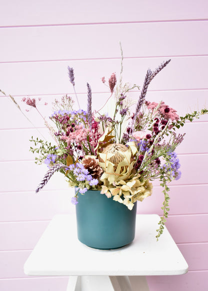 Pink background with a dried flower arrangement sitting on a white stool. A teal green ceramic pot with sage green, lilac, burgundy and lavender everlasting and dried flowers