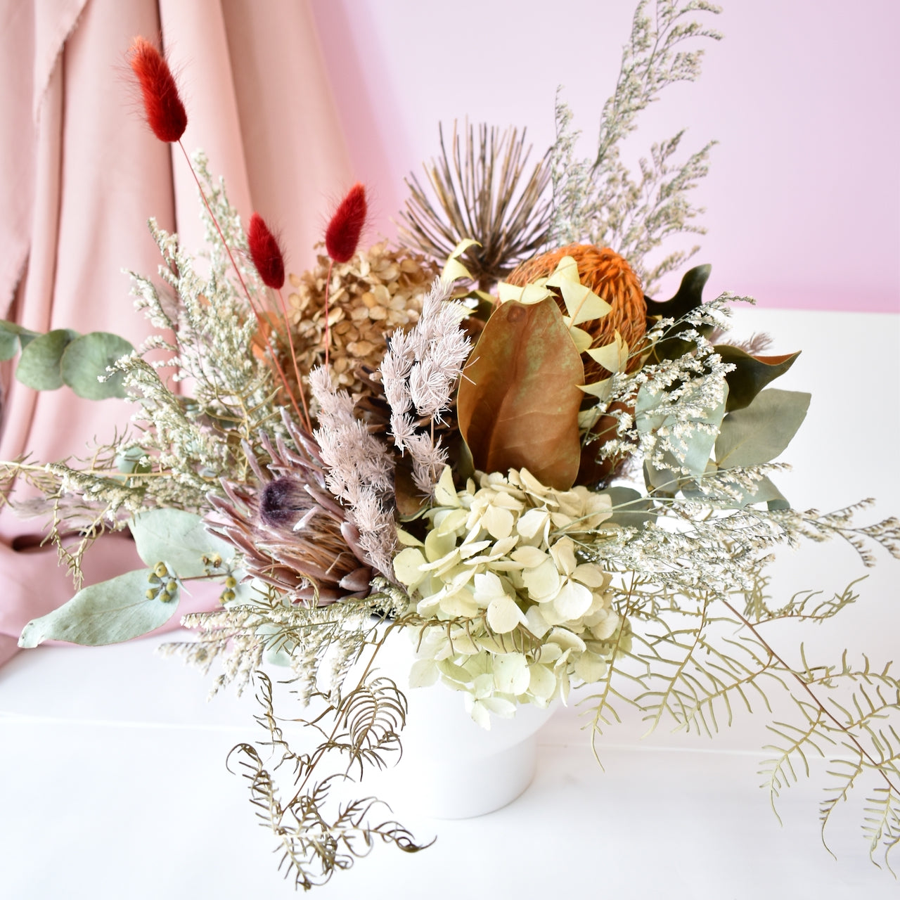 A dried flower arrangement, red bunny tails, magnolia foliage, protea, banksia, hydrangea and fern. Natural earthy colours.