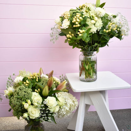 A pink background with two vases of white flowers sitting in front of. One vase sitting on white stool. White flower bouquet in glass vase