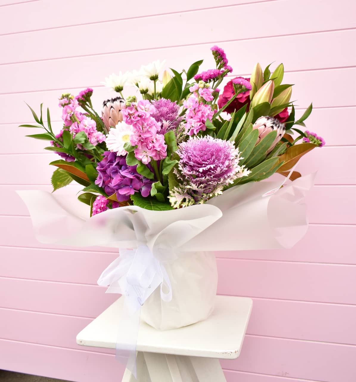 White vase with large purple and green flower arrangement against a pink back ground