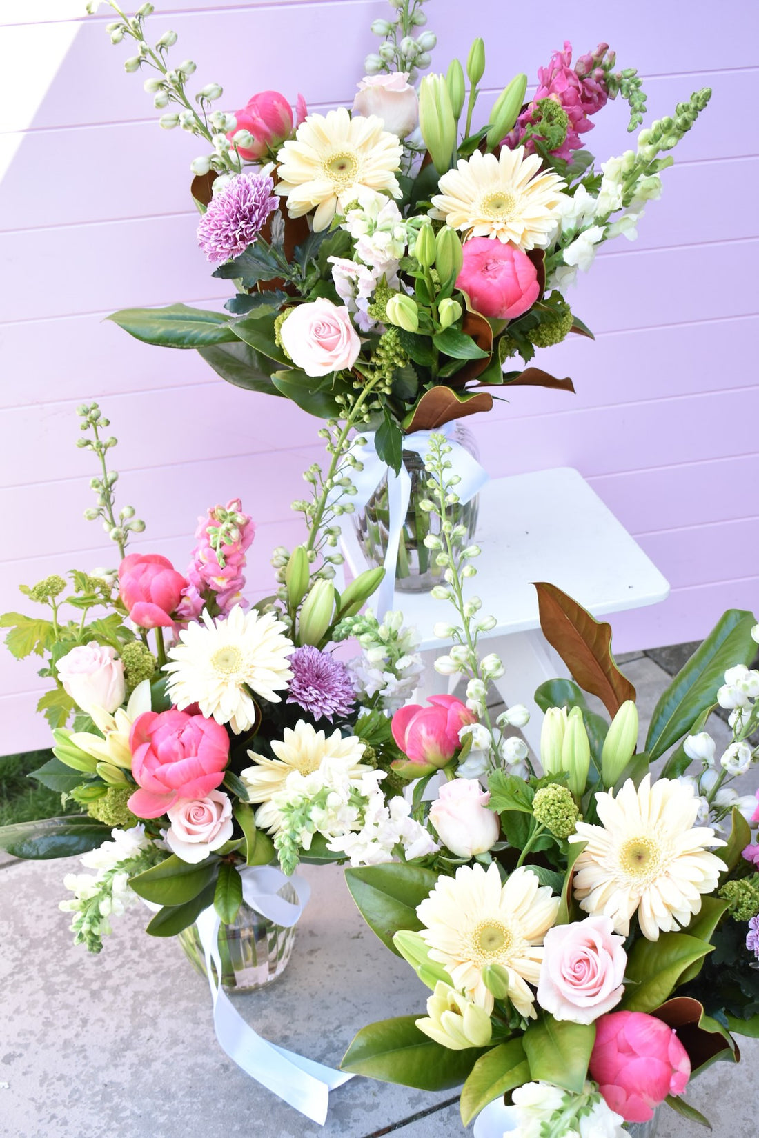 A pink background with two vases of white flowers sitting in front of. One vase sitting on white stool. Pretty flower bouquet in glass vase in colours of pink, peach, purple, white and green. Including peonies and roses