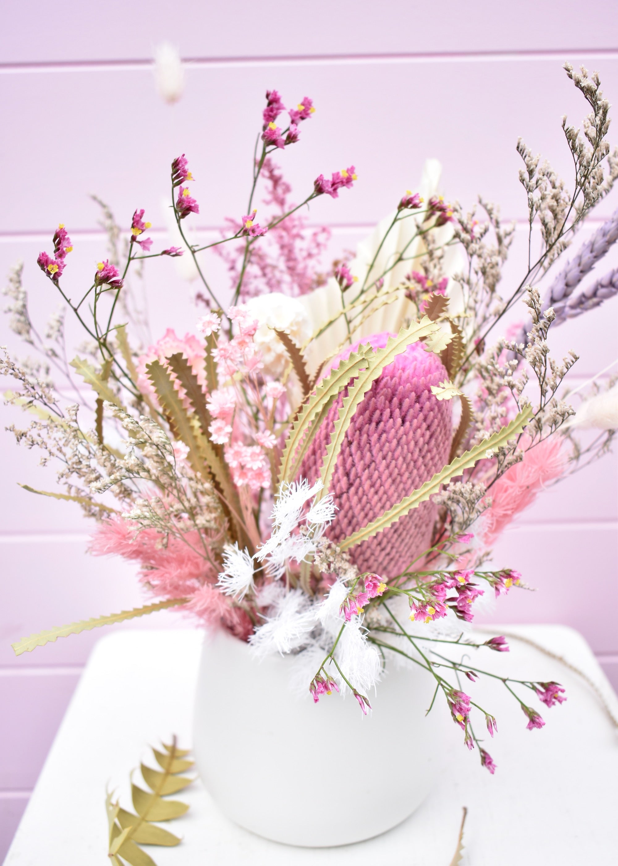 Dried flower arrangement in a white ceramic pot. Pink and white everlasting flowers, banksia, wheat, misty, ming fern, bunny tails and more. Sitting on a white stool in front of a pale pink backdrop.