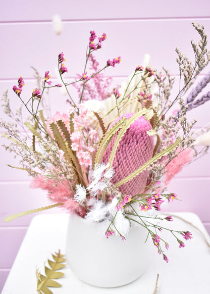 Dried flower arrangement in a white ceramic pot. Pink and white everlasting flowers, banksia, wheat, misty, ming fern, bunny tails and more. Sitting on a white stool in front of a pale pink backdrop.