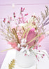 Dried flower arrangement in a white ceramic pot. Pink and white everlasting flowers, banksia, wheat, misty, ming fern, bunny tails and more. Sitting on a white stool in front of a pale pink backdrop.