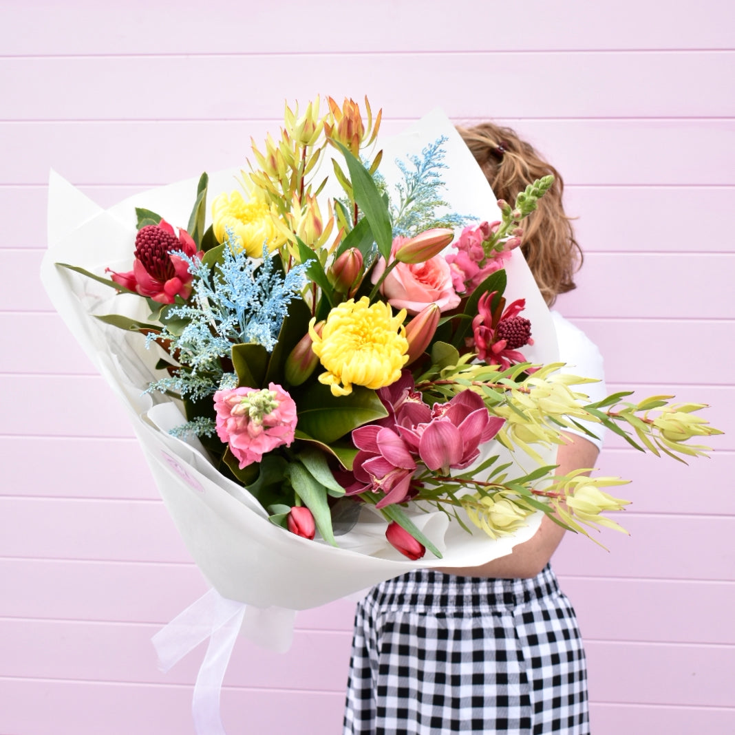 A female florist standing in front of a pink background, holding a large colourful fresh flower bouquet