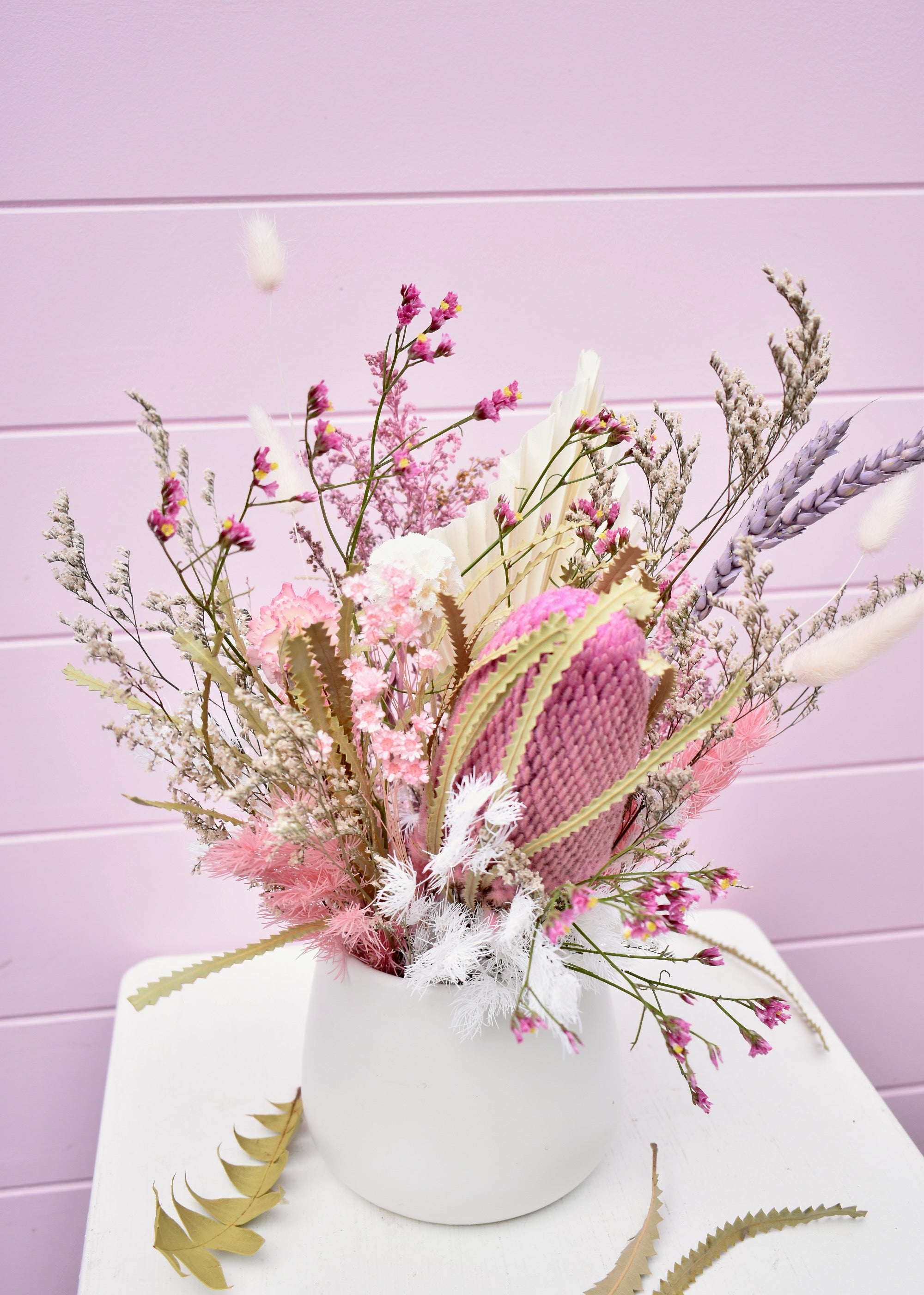 Dried flower arrangement in a white ceramic pot. Pink and white everlasting flowers, banksia, wheat, misty, ming fern, bunny tails and more. Sitting on a white stool in front of a pale pink backdrop.