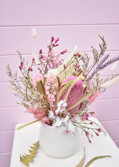 Dried flower arrangement in a white ceramic pot. Pink and white everlasting flowers, banksia, wheat, misty, ming fern, bunny tails and more. Sitting on a white stool in front of a pale pink backdrop.