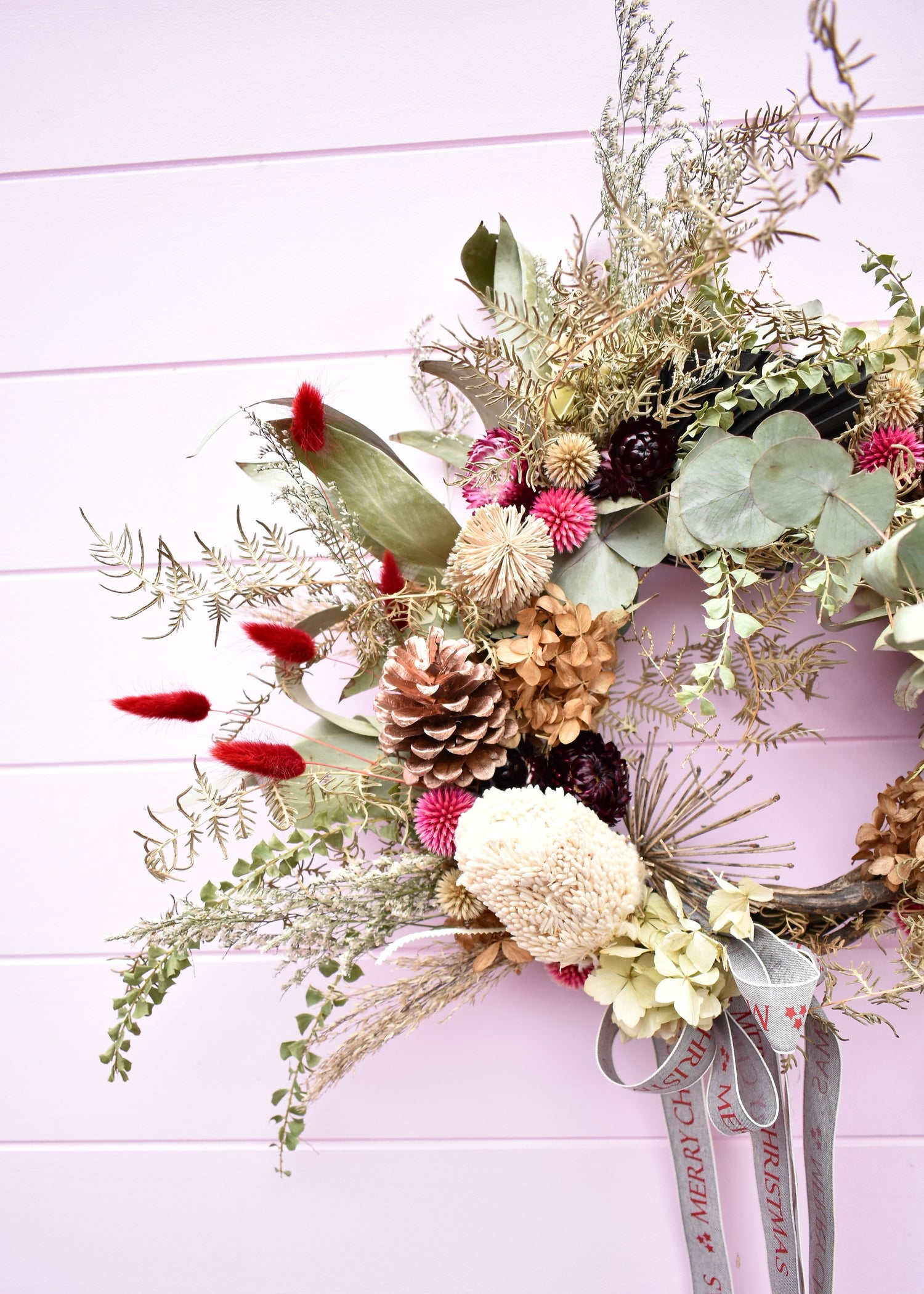 A Christmas wreath made with dried and everlasting flowers. Banksia, eucalyptus, strawflowers, fern and Christmas ribbon. Made by a Florist and on a pink background.