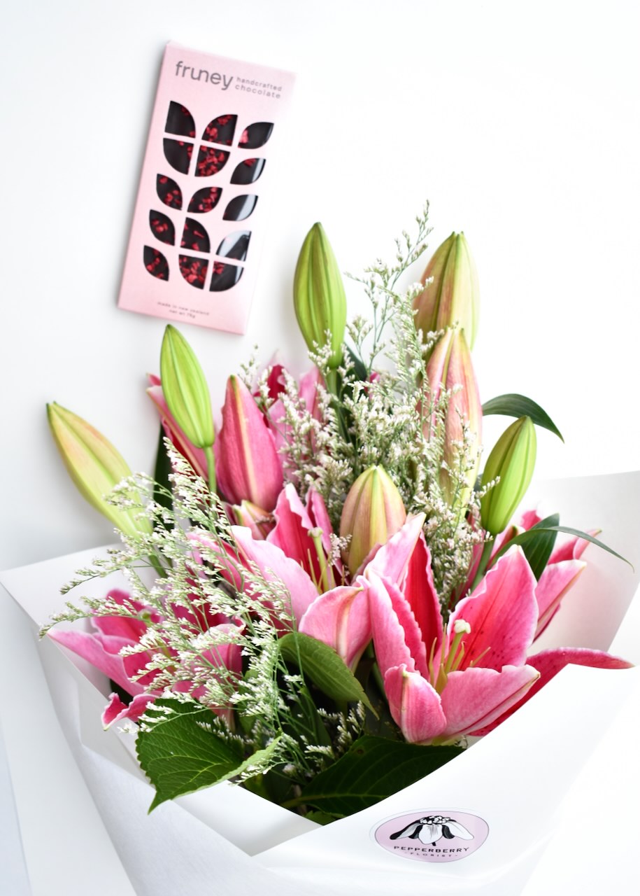 Pretty pink oriental lilies with soft white filler flower. Wrapped in white paper alongside a bar of dark chocolate &amp; raspberry in pink packaging.