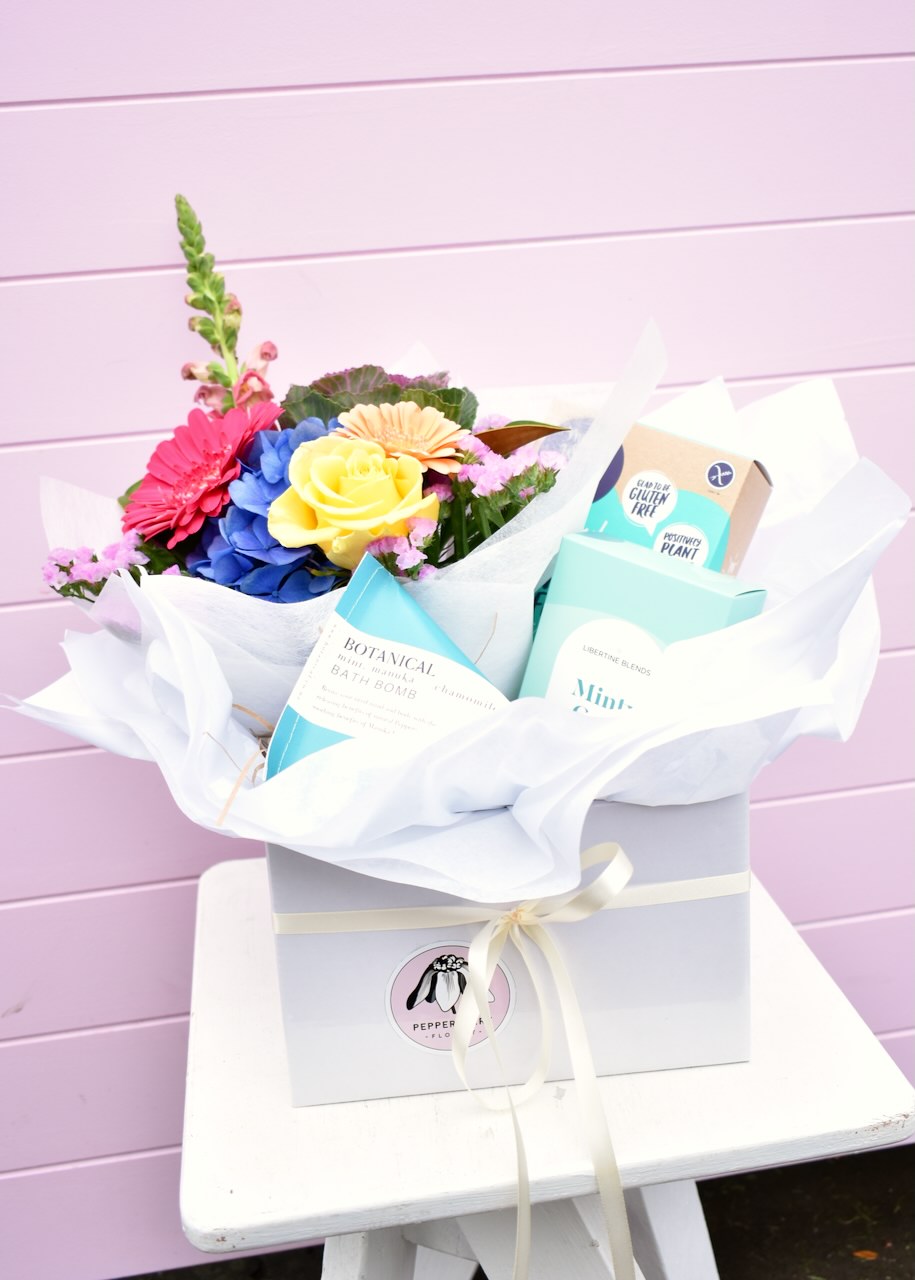 Florists gift box sitting on a white stool in front of a pink backdrop. White box containg products made in New Zealand. Fresh flower bouquet, herbal tea, artisan cookies and a bath bomb. 