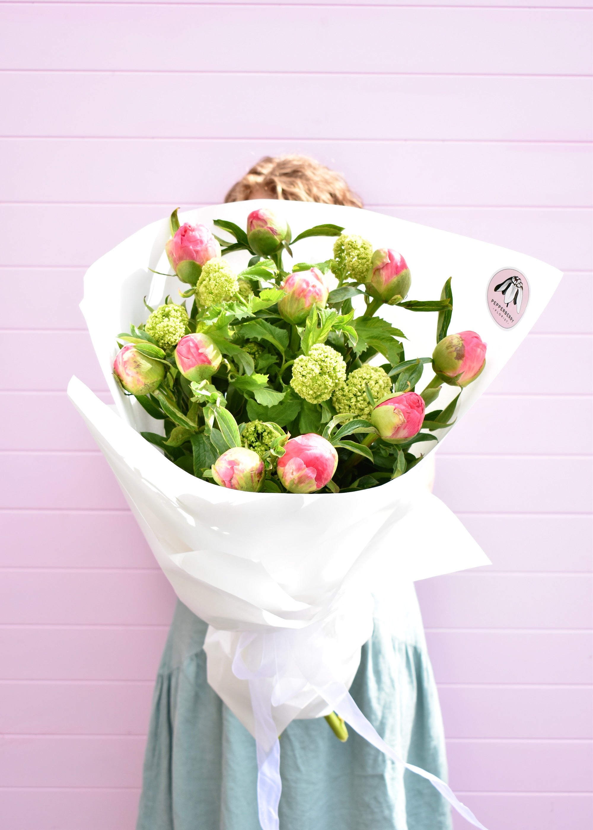 A florist holding a big bunch of coral charm peony roses, gift wrapped in white paper with Pepperberry Florist logo