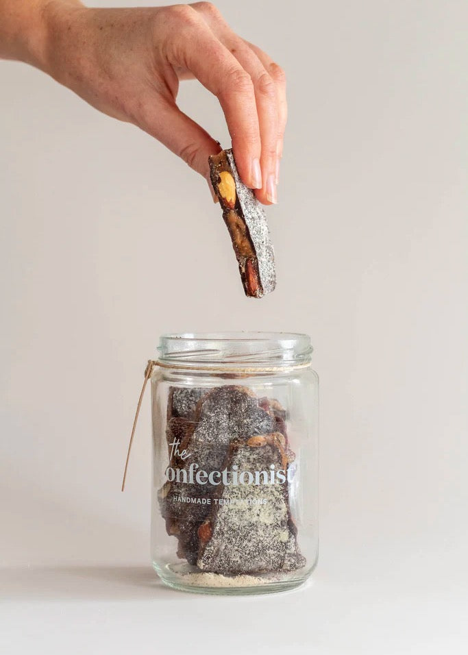 A product shot of a hand reachong into a jar of dark chocolate and almond toffee