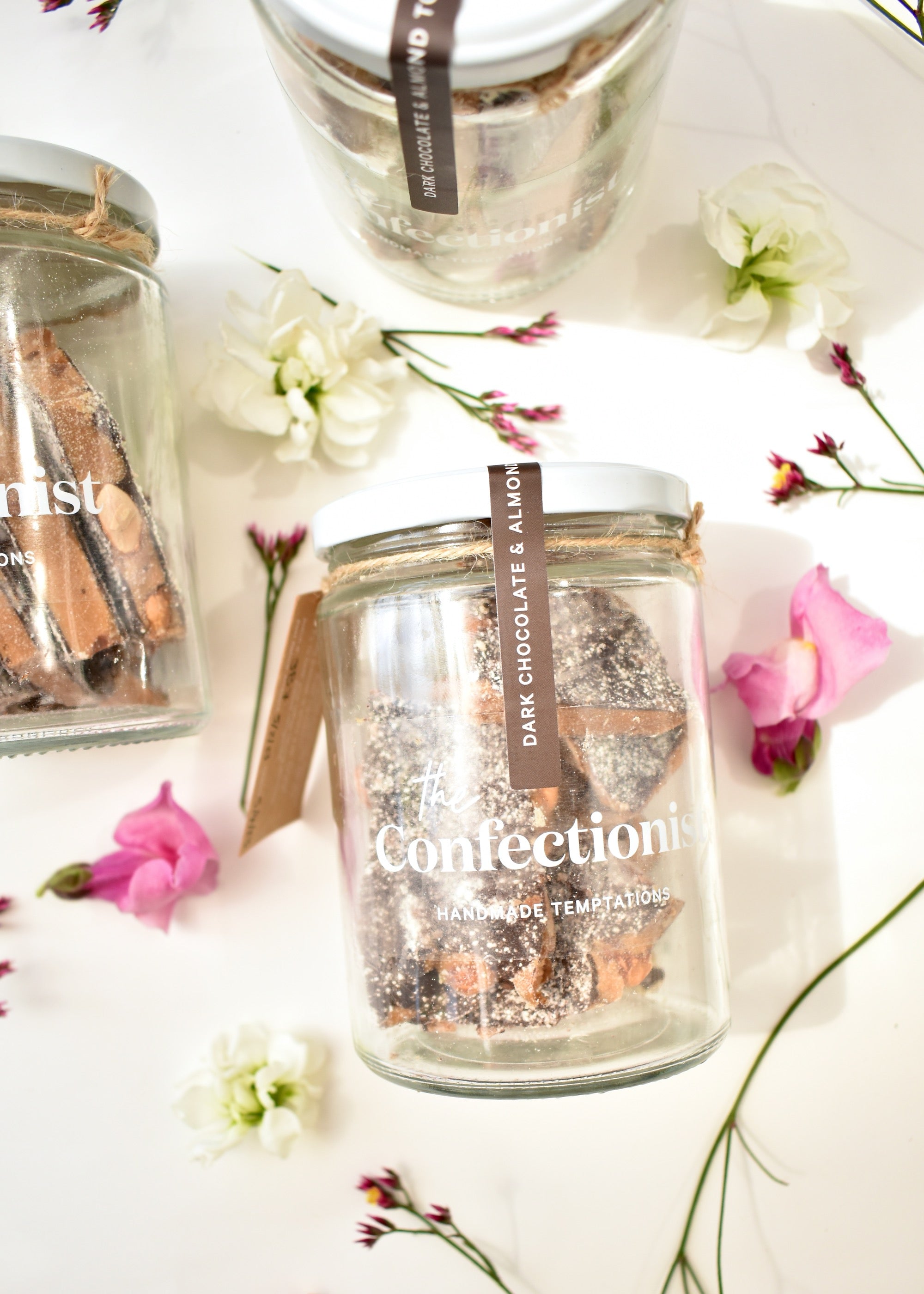 Flat lay image of jars of dark chocolate and almond toffee. White background with pink and white flowers