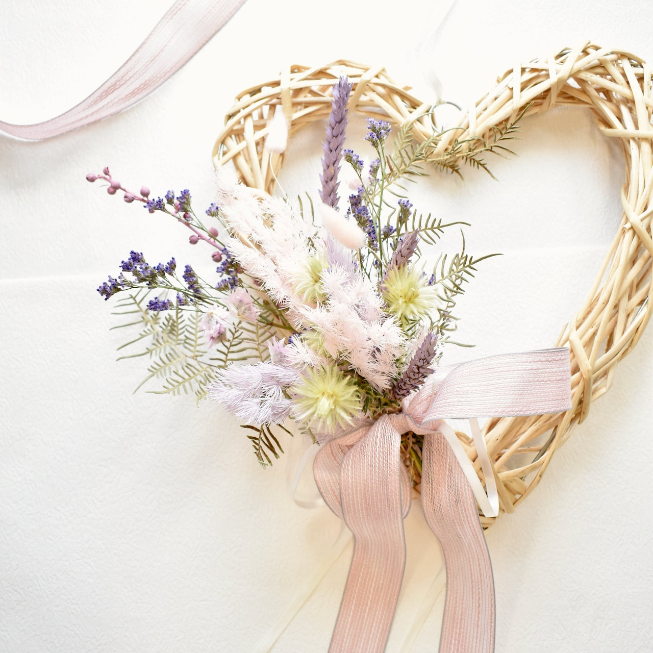 A cream wicker heart wreath on a pink backdrop, with a posy of dried flowers in white, cream, pink and purple with pale pink ribbon